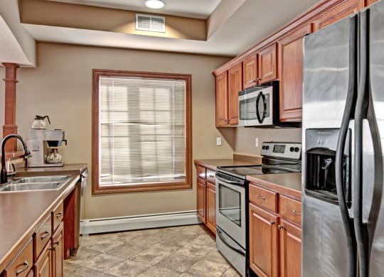 A modern kitchen here at Grand River Estates Apartment Homes with wooden cabinets, stainless steel appliances, and tiled floor. Sunlight streams through a window, creating a warm, inviting ambiance.