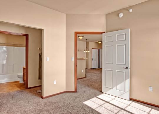 Vacant apartment interior here at Grand River Estates Apartment with beige walls and carpet. Open door reveals dining area with pendant lights. Bathroom entrance on the left, creating a warm, inviting feel.