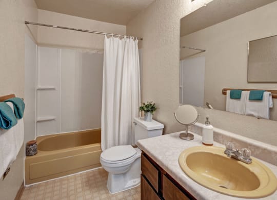Bathroom here at Greenbriar Apartments with a yellow bathtub and sink, white shower curtain, and countertop. Towels and a small plant add color. Clean and neutral decor.