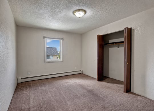 Bedroom here at Greenbriar Apartments with beige walls and brown carpet. A single window shows a roof and sky. Open closet with wooden doors on the right. Simple and neutral tone.