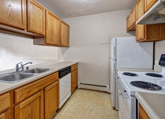 Narrow kitchen here at Greenbriar Apartments with wooden cabinets, beige countertops, and appliances including a stainless-steel sink, dishwasher, white stove, and fridge. Cozy and functional.