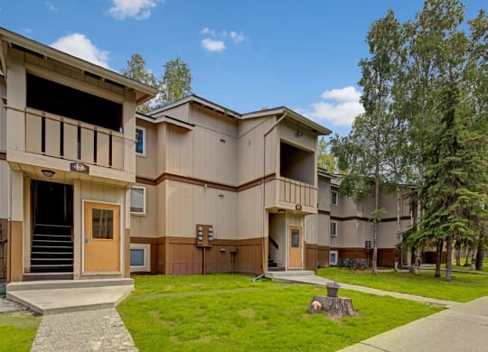 Two-story beige apartment buildings here at Greenbriar Apartments with brown trim amidst a landscaped, grassy area with trees and blue sky, conveying tranquility.
