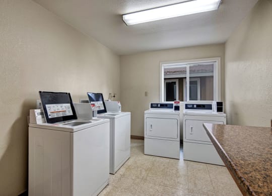 A clean, brightly-lit laundry room here at Greenbriar Apartments with four white washing machines and dryers lined against cream-colored walls. A window reflects natural light.