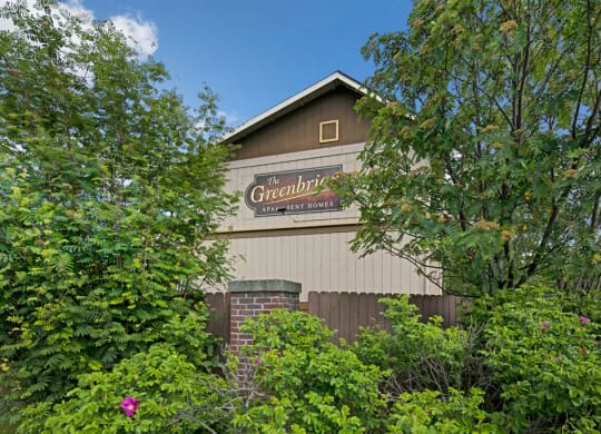 A building sign for "The Greenbriar Apartment Homes" is partially obscured by lush green trees and shrubs, with a clear blue sky in the background.