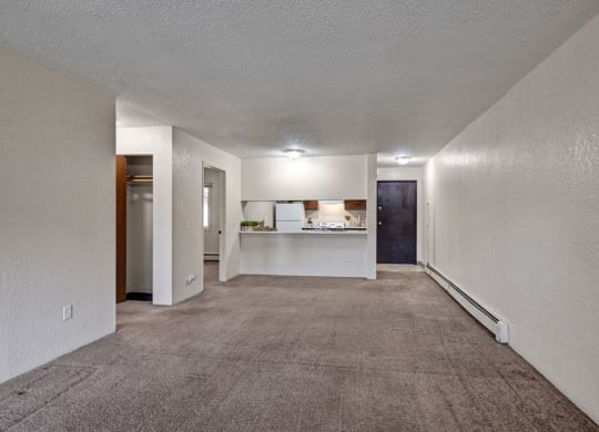 Empty living room here at Greenbriar Apartments with beige carpet and white walls, featuring a small closet on the left, a kitchen with a counter in the middle, and a dark wooden door on the right. Bright overhead lighting creates a clean, spacious feel.
