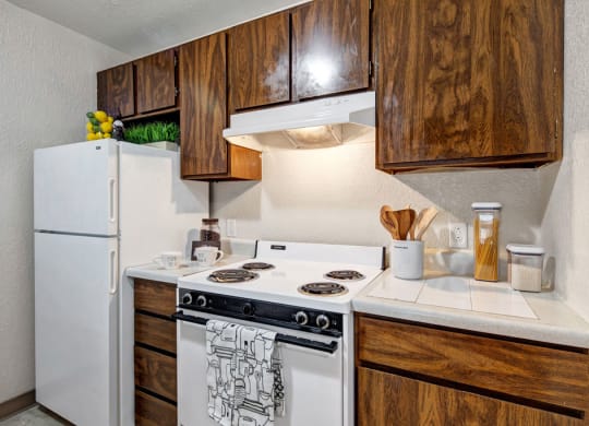 A compact kitchen here at Greenbriar Apartments with wooden cabinets, white appliances, and decor such as lemons, plants, and utensils. It's cozy and organized.