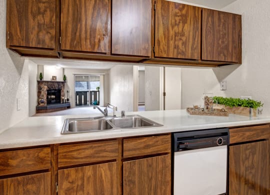 Stylish kitchen here at Greenbriar Apartments with wooden cabinets, double sink, and dishwasher. A pass-through window reveals a cozy living room with a fireplace, creating a warm tone.