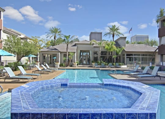 Luxurious pool area here at The Hawthorne Apartments with blue jacuzzi in the foreground, surrounded by lounge chairs. Palm trees and a modern building create a serene, sunny atmosphere.