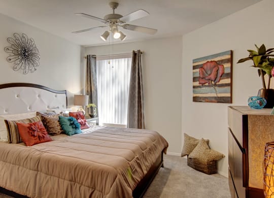 Bedroom here at The Hawthorne Apartments with a neutral tone features a large bed with colorful pillows. A flower painting and metal wall art add warmth, while soft light from a lamp enhances coziness.