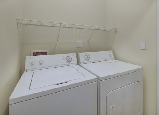 A laundry room her at The Highlands Luxury Apartments with a white washer and dryer side by side. Above them is a wire shelf. The walls are light beige, creating a clean, tidy feel.