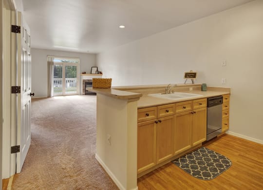Open-plan living space here at The Highlands Luxury Apartments with light wood kitchen cabinets, a sink, and dishwasher. Cozy carpeted area leads to a sunlit balcony, creating an inviting ambiance.