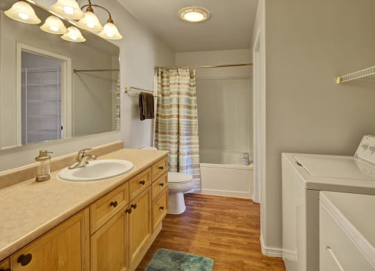 Cozy bathroom here at The Highlands Luxury Apartments with a wooden vanity, oval sink, and mirror. Bathtub with a striped curtain, towel rack, washer, and wood floor. Bright and clean.
