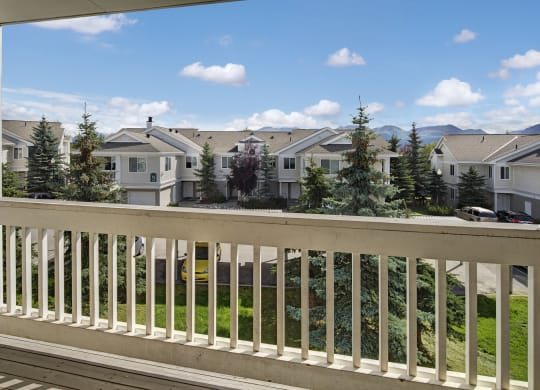 View from a balcony here at The Highlands Luxury Apartments overlooking a row of townhouses with a bright blue sky and scattered clouds. Tall pine trees and parked cars line the street below.