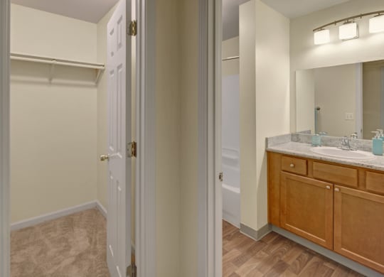 A modern bathroom here at Hillside Chalet Apartments features a spacious vanity with a marble countertop, wooden cabinets, and dual sinks. Adjacent is a carpeted closet space.
