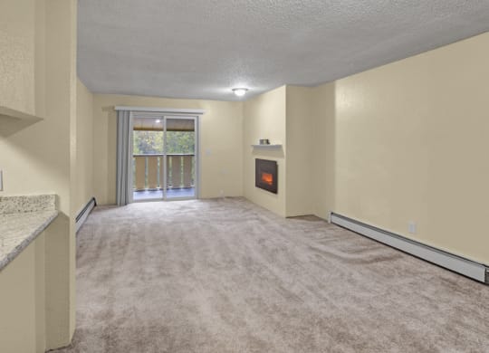 Empty living room here at Hillside Chalet Apartments with beige walls and carpet, featuring a corner electric fireplace. Sliding glass door leads to a balcony with green foliage outside.