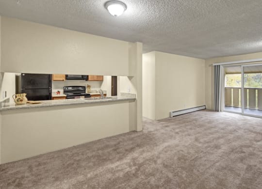 Spacious empty living room here at Hillside Chalet Apartments with beige carpet, open kitchen with light wood cabinets, and large sliding glass doors leading to a balcony with greenery.