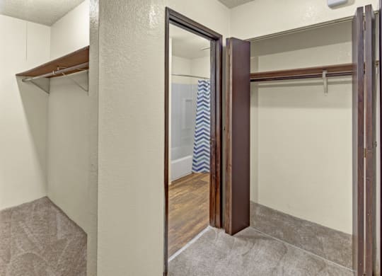 A small, empty walk-in closet here at Hillside Chalet Apartments with beige walls, open wooden shelves, and brown carpet. Leads to a bathroom with a chevron-patterned shower curtain.