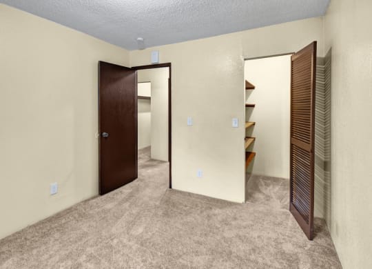 Empty beige bedroom here at Hillside Chalet Apartments with brown carpet, featuring an open door leading to another space with shelving. The atmosphere is simple and neutral.