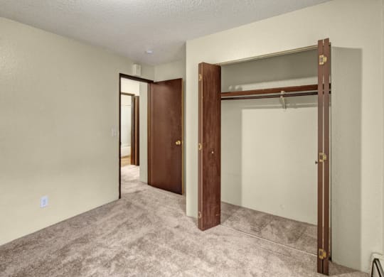 Empty bedroom here at Hillside Chalet Apartments with textured beige walls and carpeted floor. An open closet with wooden doors is on the right, and a doorway leads to a bathroom.