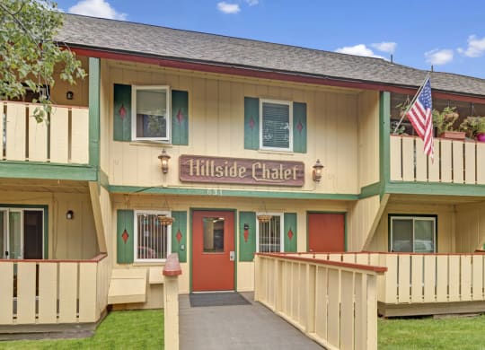 Two-story Hillside Chalet Apartment building with a rustic design, labeled "Hillside Chalet." Features balconies with potted plants, a wooden fence, and an American flag.
