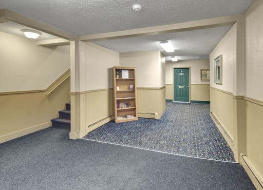 A quiet, well-lit hallway here at Hillside Chalet Apartments with cream-colored walls and patterned carpet. A green door is at the end, with stairs and a bookshelf on the left.