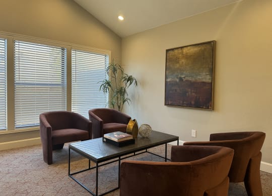 A cozy seating area here at Eagle Ridge Apartment Homes with four brown chairs around a black coffee table. A large abstract painting and a tall plant decorate the warm, well-lit room.