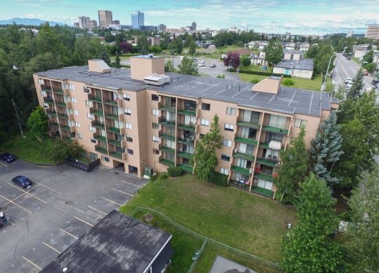 Aerial view of a beige, five-story Outlook apartment building with balconies, surrounded by green trees and a parking lot. City skyline in the distance under a cloudy blue sky.