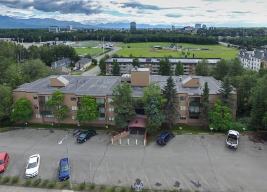 Aerial view of a brown Outlook apartment building with a parking lot in front, surrounded by trees. In the background, a sports field, trees, and a cloudy sky with distant mountains.