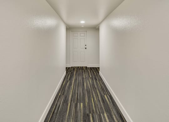 Narrow, brightly lit hallway at La Maisonnette Apartments with white walls and door at the end. The floor features dark patterned carpeting with subtle yellow stripes, creating a modern feel.