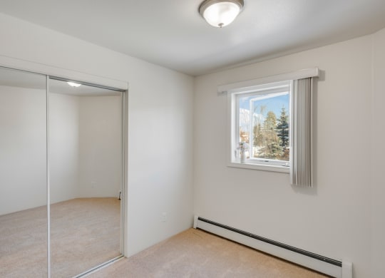 Small bedroom here at La Maisonnette Apartments with beige carpet and white walls, featuring a window with vertical blinds, a ceiling light, and a mirrored closet door reflecting light.