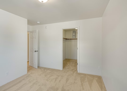 Bright bedroom here at La Maisonnette Apartments with beige carpet and white walls. Features an open closet with a "HOME" sign, and an ajar door leading to another room. Calm atmosphere.