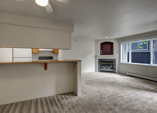 Spacious living area here at La Maisonnette Apartments with beige carpet, a white ceiling fan, and natural light from a large window. Features a fireplace and open kitchen with a counter.