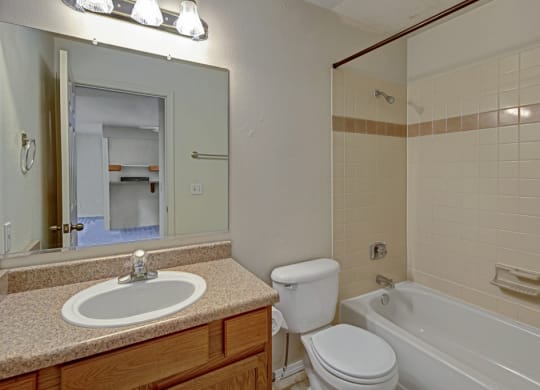 A bathroom here at La Maisonnette Apartments with beige tiled walls features a bathtub, a white toilet, and a brown vanity with a sink. A mirror reflects a doorway to another room.