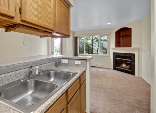 Compact kitchen here at La Maisonnette Apartments with oak cabinets, gray countertops, and stainless steel sink. Open view to living room with carpet, large window, and lit fireplace. Cozy ambiance.