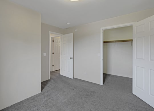 Empty bedroom here at La Maisonnette Apartments with gray carpet and light beige walls. Two open doors reveal a closet and a hallway. The space feels clean and minimalistic.