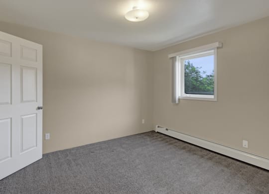 Bedroom here at La Maisonnette Apartments with beige walls, a gray carpet, and a white door ajar. A window with blinds lets natural light in, creating a calm, neutral ambiance.