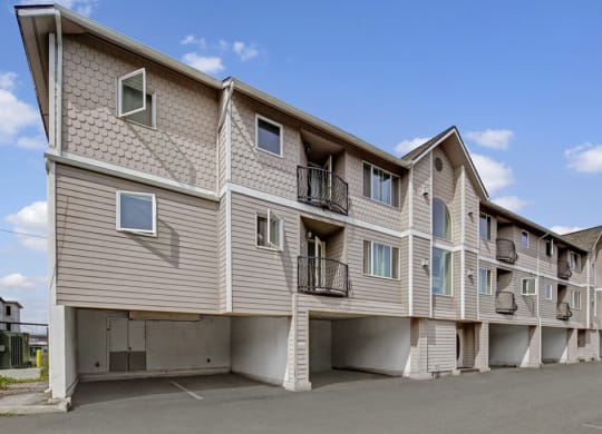 Three-story La Maisonnette apartment building with beige siding, decorative shingles, and small balconies. Open parking space underneath. Clear blue sky with a few clouds.