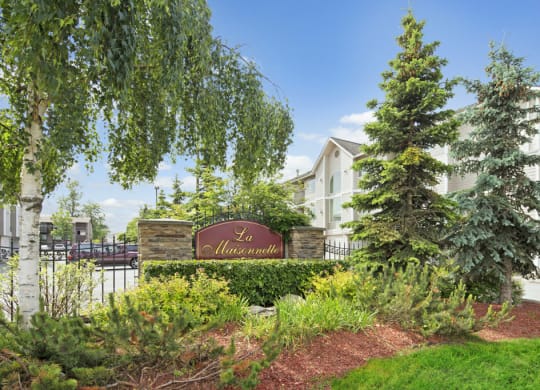 Lush garden with trees surrounds a sign labeled "La Maisonnette." A light-colored building is in the background under a clear blue sky.