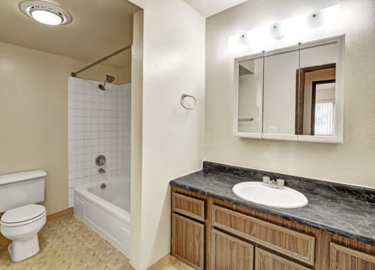 A tidy bathroom here at Ladera Villas Apartments with a bathtub-shower combo, white tiles, and a white toilet. A wooden vanity with a marble countertop sits beneath a lit mirror.