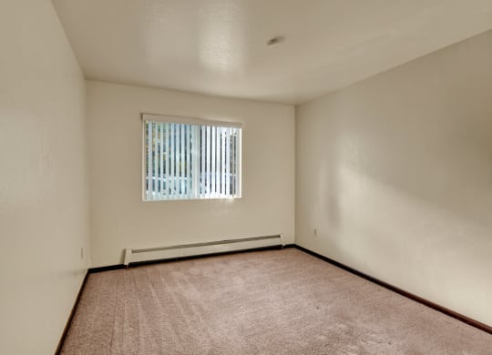 Bedroom here at Ladera Villas Apartments with beige carpet and cream walls, lit by sunlight through vertical blinds on a window. The tone is warm and inviting.