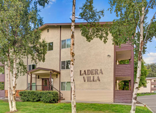 Three-story beige Ladera Villa apartment building labeled "Ladera Villa" with brown accents. Surrounded by lush green trees and grass under a clear blue sky.
