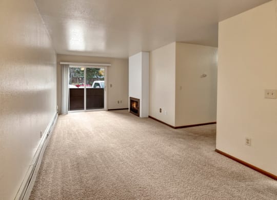 A spacious, empty living room here at Ladera Villas Apartments with beige carpet and white walls. A lit fireplace adds warmth. A sliding glass door opens to a patio, revealing parked cars outside.