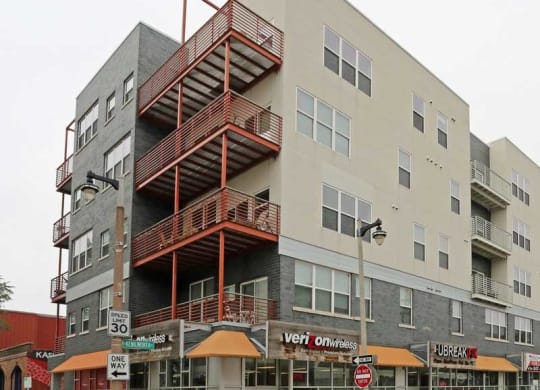 Street view of the Latitude Apartments, a modern, multi-story apartment building with retail stores on the ground floor. Orange awnings and metal balconies create a contemporary feel.