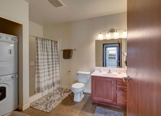 Modern bathroom at the Latitude Apartments with light wood cabinetry, a large mirror, and warm lighting. A striped shower curtain is adjacent to a stacked washer and dryer.