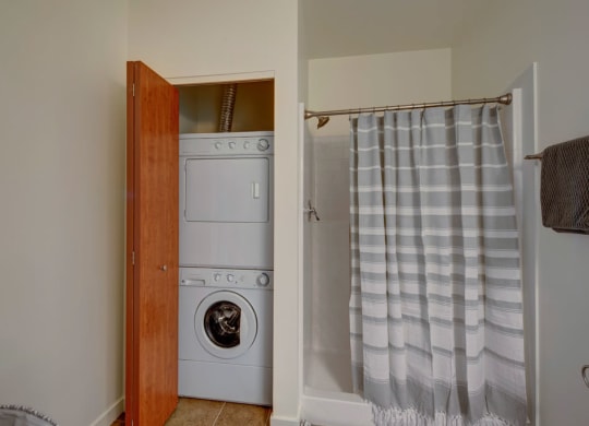 The Latitude Apartments bathroom with a striped shower curtain concealing a shower area. A stackable washer and dryer are inside an open wooden closet.