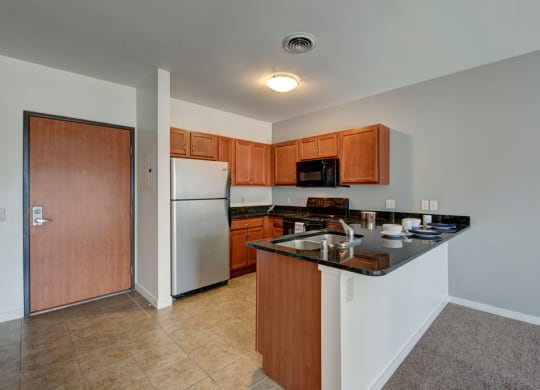 Modern kitchen at Latitude Apartments with stainless steel fridge, wooden cabinets, and a black granite countertop. Includes a built-in sink, microwave, and oven. Warm, inviting tone.