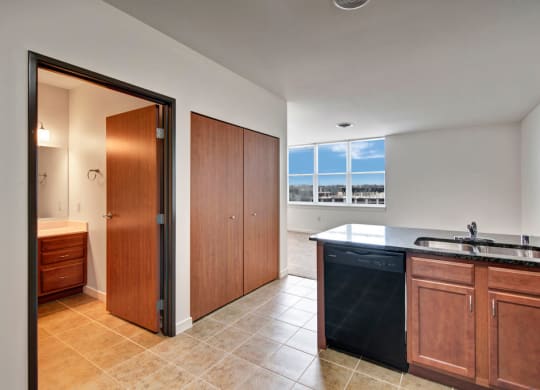 Modern apartment interior with open kitchen, featuring wooden cabinets, black counters, and tile floor. Bright window view and a closed bathroom door. The Latitude Apartments.