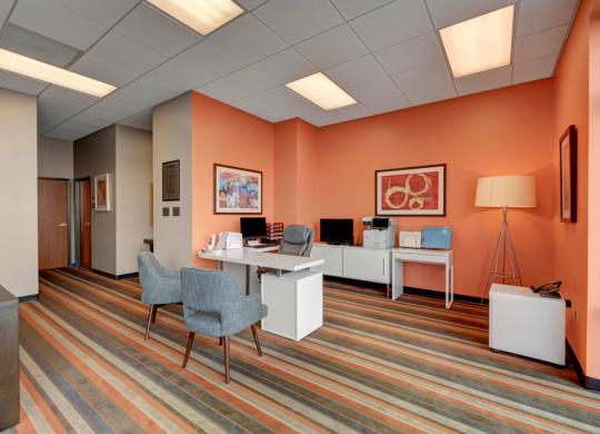 A modern business center with peach walls and striped carpet. Features a white desk with a computer and gray chairs. Artwork and a tall lamp add warmth. The Latitude Apartments.