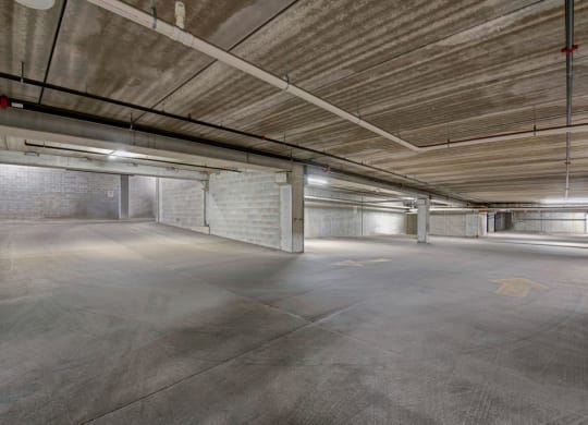 Empty underground parking garage at the Latitude Apartments with concrete walls, ceiling, and floor. Bright fluorescent lights illuminate the space, creating a stark, industrial feel. Yellow arrows mark directions on the floor.