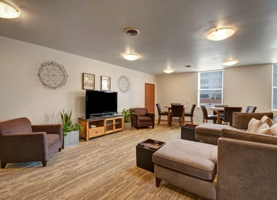 Spacious resident lounge at Latitude Apartments with brown couches, a TV, and geometric wall art. Dining tables line the back wall under large windows, creating a cozy, inviting atmosphere.